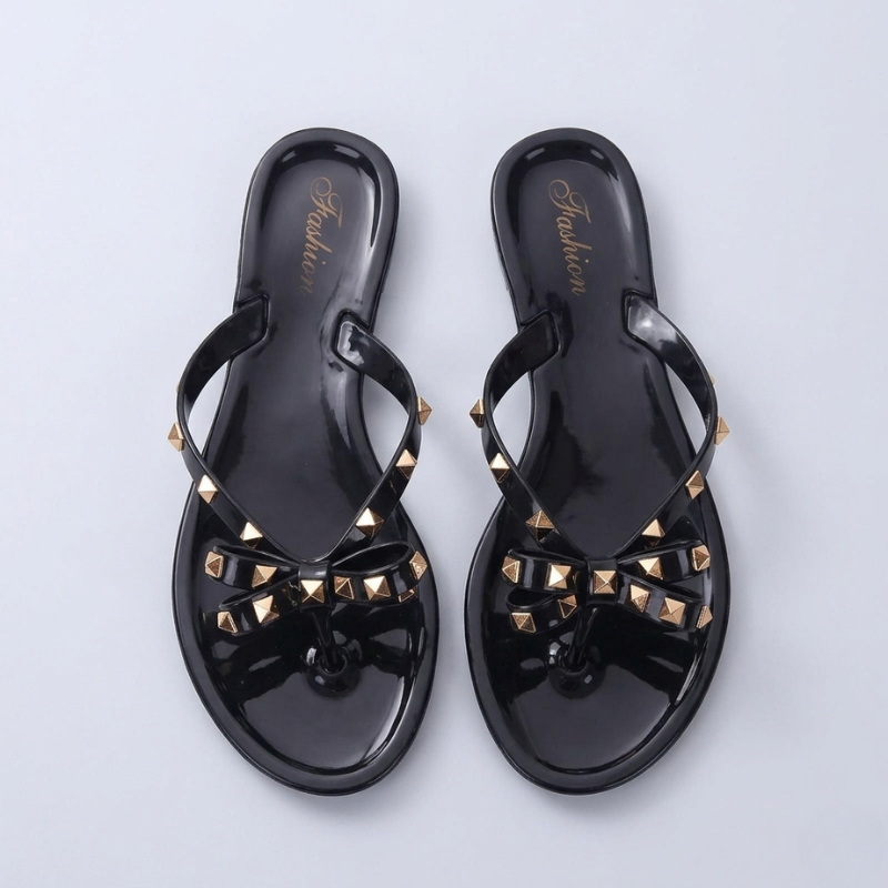 A pair of black jelly flip flops with gold studded bows shown from a top-down perspective on a light grey background.
