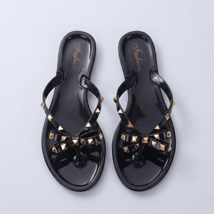 A pair of black jelly flip flops with gold studded bows shown from a top-down perspective on a light grey background.