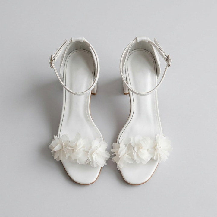 Top-down view of a pair of white ankle-strap block heels with chiffon floral details on a neutral background.