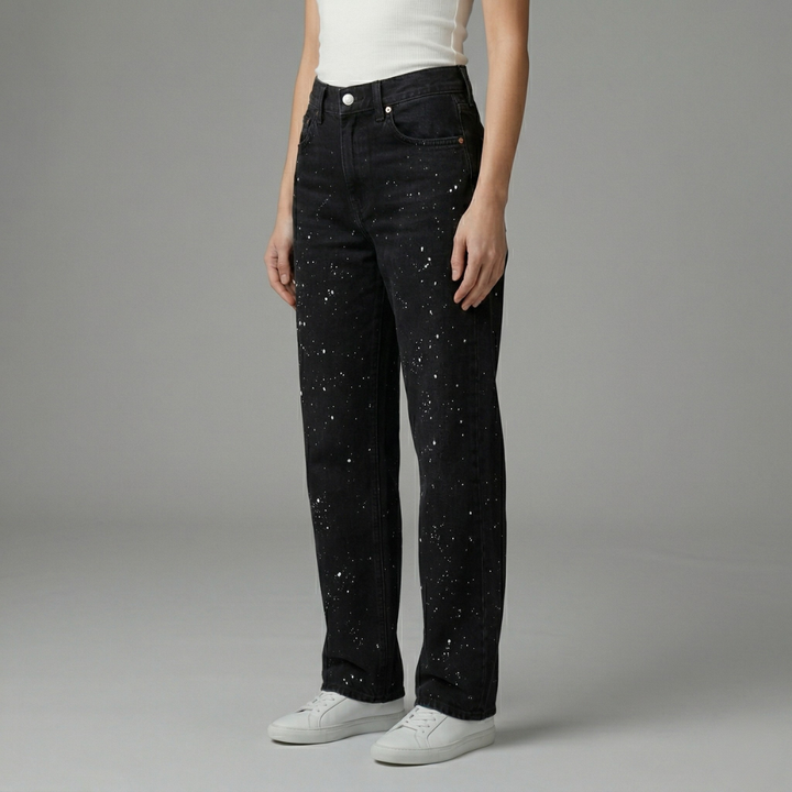 Front view of black denim straight-leg jeans featuring white paint splatter accents on a female model.