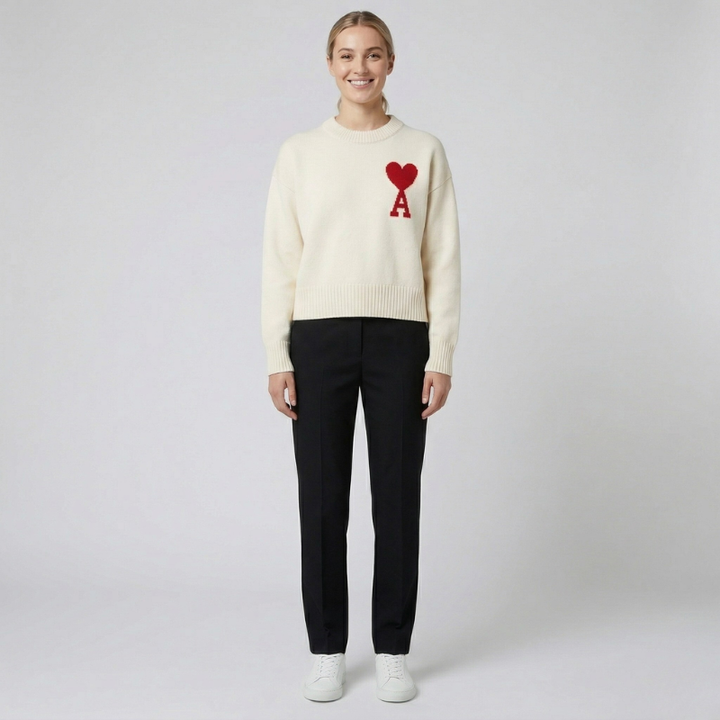 Minimalist studio shot of a cream-colored sweater with a red heart logo paired with black trousers.