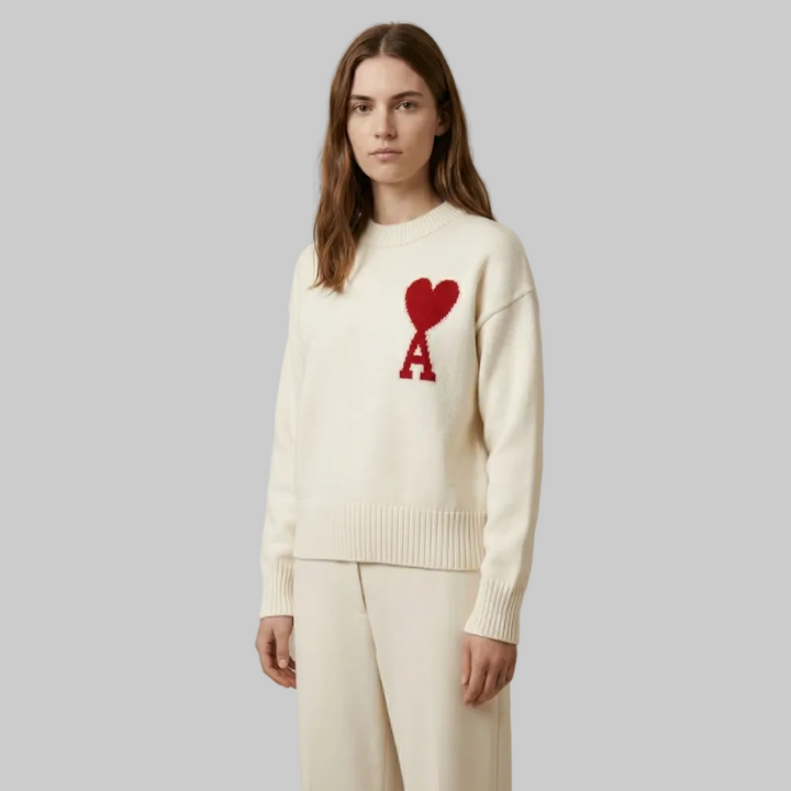 Front view of a woman wearing a cream knit sweater with a red heart emblem, paired with matching cream pants.