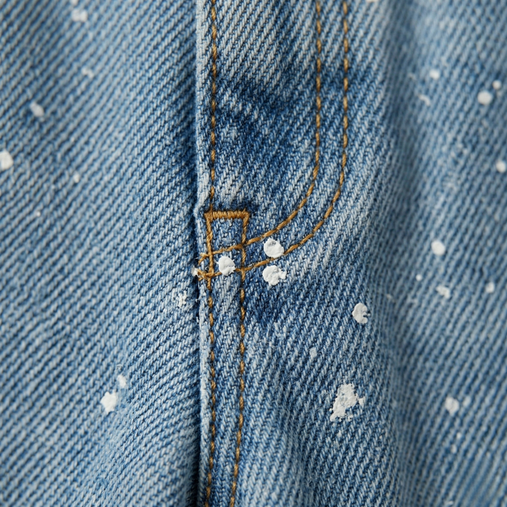 Extreme close-up of the denim texture, yellow contrast stitching, and white paint splatter details near the fly.