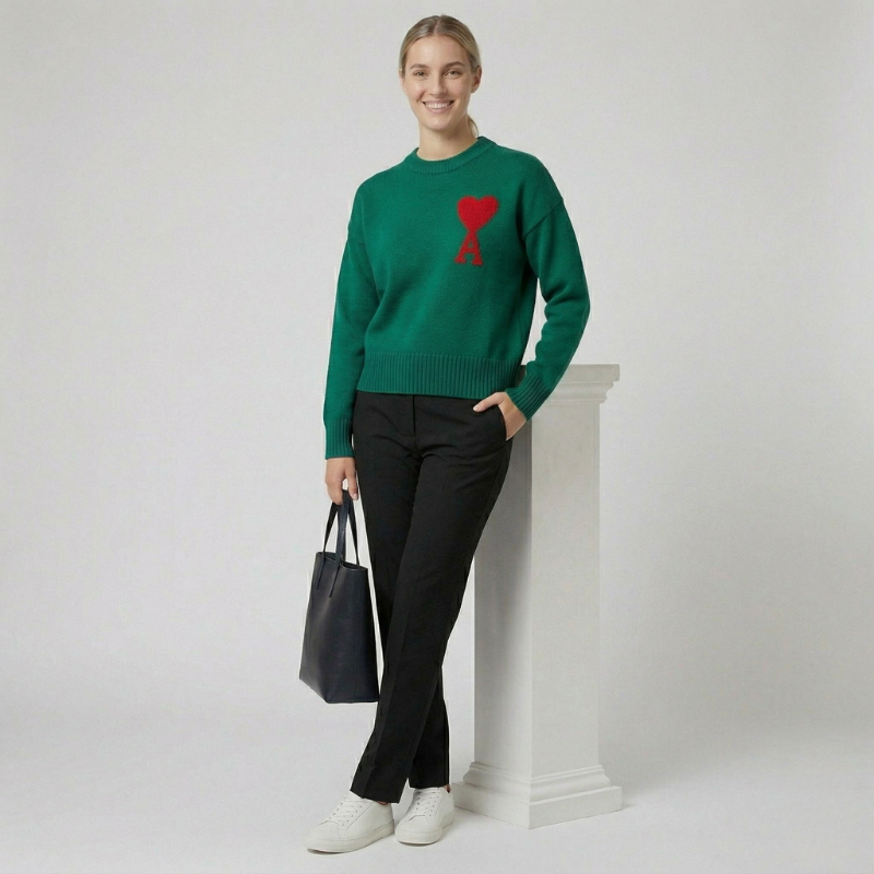 A woman posing in a forest green crewneck sweater with a red heart logo and black pants.