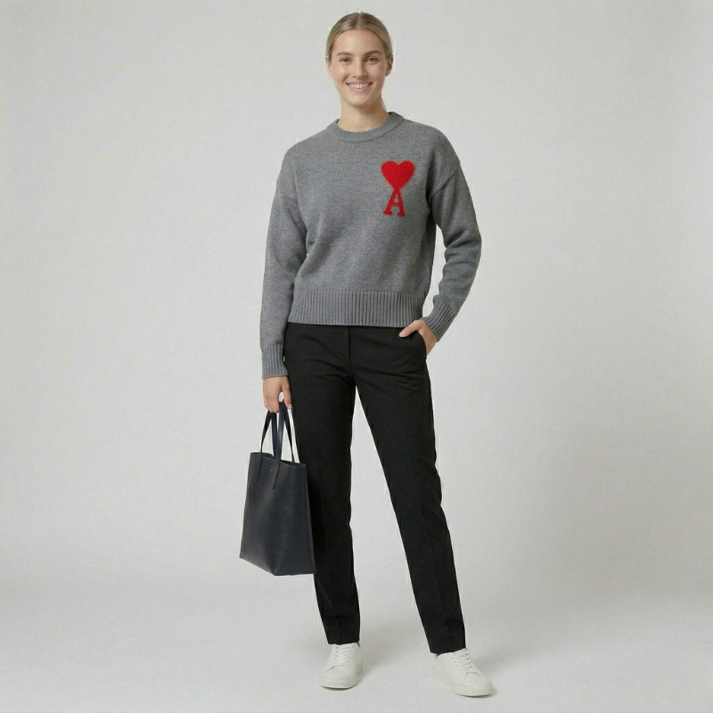 A woman wearing a heather gray crewneck sweater with a red heart logo, styled with black trousers and a handbag.